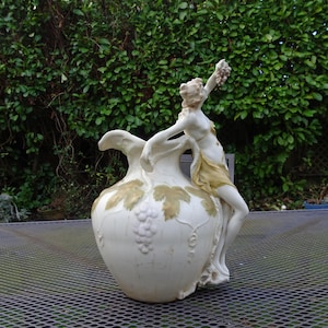May include: A cream-colored ceramic pitcher featuring a sculpted figure draped in a gold-toned cloth, reaching upward toward a cluster of grapes. The body of the vessel is decorated with embossed grape clusters and vine leaves in a muted gold finish.