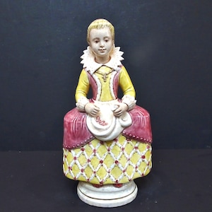 以下が含まれることがあります： 歴史的なドレスを着た、座っている女性の陶器製の置物。このフィギュアは、ピンクのアクセントが付いた黄色のボディス、白いフリルカラー、ピンクのスカートを着用しています。スカートは黄色と白のダイヤモンド柄です。この置物は白い台座の上にあります。