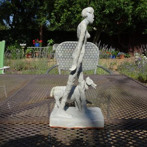 Puede incluir: Una escultura de color gris claro que representa a una figura de pie caminando junto a un perro pequeño. La pieza está elaborada con un acabado mate y texturizado, montada sobre una base rectangular. Las figuras tienen una forma estilizada y alargada.