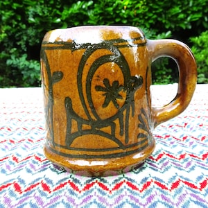 Peut inclure: Une tasse en céramique artisanale avec un glaçage marron et des motifs abstraits vert foncé. La tasse a une grande anse et est posée sur une nappe colorée à motifs. La tasse mesure environ 10 cm de haut.