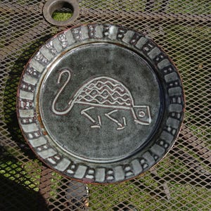 Puede incluir: Un plato de cerámica con un centro gris oscuro y un borde decorativo. El plato presenta un diseño de tortuga blanca en el centro. El borde tiene un patrón repetitivo de formas rectangulares. El plato está sobre una superficie metálica.