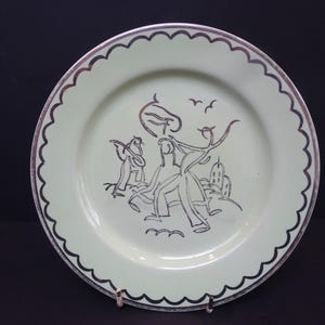 May include: A light green plate with a scalloped edge and a silver rim. The plate features a black and silver illustration of figures and birds. A decorative piece for display.