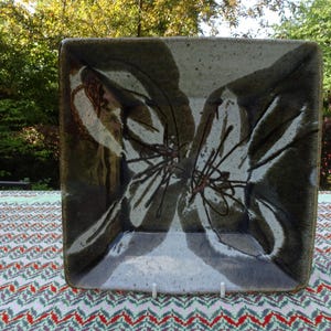 Può includere: Piatto quadrato in ceramica con un design unico. Il piatto presenta un motivo floreale marrone scuro e bianco. Il piatto è esposto su una tovaglia fantasia, con uno sfondo naturale all'aperto.