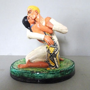 Puede incluir: Una figura de cerámica que representa a un hombre y una mujer abrazándose. El hombre, con cabello rubio y camisa blanca, está arrodillado mientras sostiene a la mujer. La mujer tiene cabello negro y una falda estampada con diseños amarillos, negros y blancos. La base es verde.