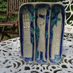 Puede incluir: Arte mural rectangular de cerámica con una combinación de colores azul, verde y blanco. La obra presenta tres paneles verticales con figuras estilizadas y elementos arquitectónicos. Expuesta sobre una mesa blanca ornamentada.