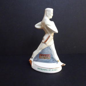 Puede incluir: Una figura de cerámica blanca de un atleta estilizado en pose de carrera. La figura tiene detalles dorados y una base azul. La base dice "XVI OLIMPIADE ROMA".