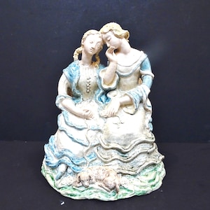 以下が含まれることがあります： 歴史的なドレスを着た2人の女性を描いた陶器製の置物。女性はブロンドの髪で、青いアクセントの白いドレスを着ています。小さな犬が足元に、緑と白の台座の上にいます。この置物はヴィンテージ感があります。