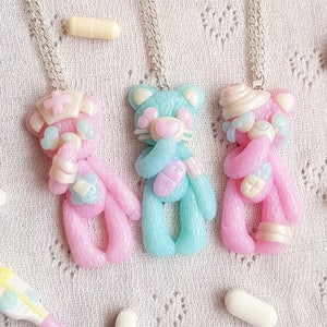 Three polymer clay necklaces depicting pastel teddy bears dressed as nurses and hospital patients. In the background a fake termomether and another hospital themed necklace. Loose pills scattered around.