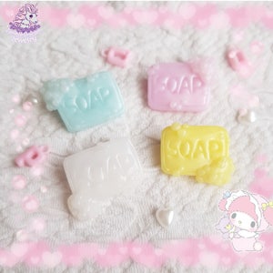 May include: Four pastel-colored soap-shaped charms with the word "SOAP" embossed on them. The charms are light blue, pink, white, and yellow, with bubbly foam details. The charms are on a white, textured surface.