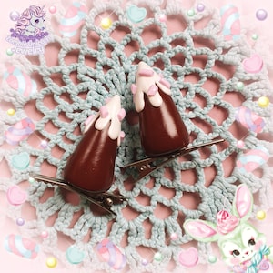 Puede incluir: Dos pinzas para el pelo marrones con forma de conos de helado con glaseado blanco y rosa. Las pinzas están sobre una base de metal plateado. El fondo es un tapete de ganchillo azul claro con caramelos pastel y un conejito de dibujos animados.