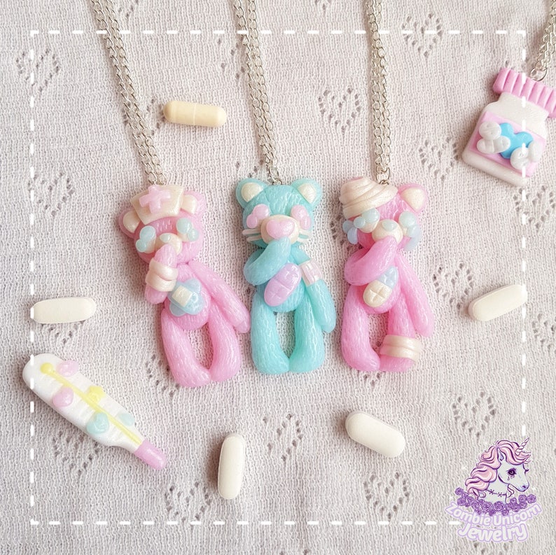 Three polymer clay necklaces depicting pastel teddy bears dressed as nurses and hospital patients. In the background a fake termomether and another hospital themed necklace. Loose pills scattered around.