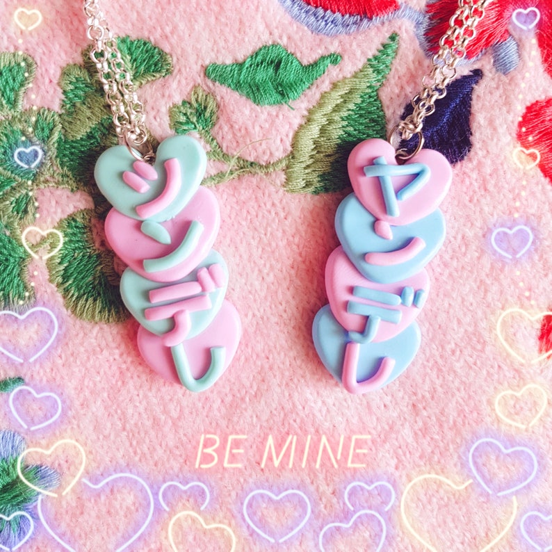 May include: Two heart-shaped pendant necklaces with Japanese characters, in pastel pink and blue, hang on silver chains. The necklaces are stacked hearts. The background is a pink, embroidered fabric with the words "BE MINE" in neon.