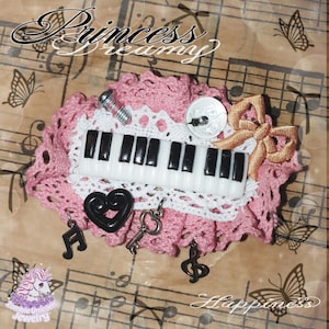 Può includere: Spilla decorativa a tema musicale con una tastiera di pianoforte in miniatura, un supporto in pizzo rosa e bianco e vari ciondoli, tra cui un cuore, una chiave e note musicali. La spilla è adornata da un fiocco e dal testo "Princess Dreamy".