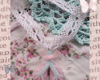 May include: A handmade necklace featuring a pink and blue lung-shaped pendant with a pink bow, suspended from a white lace ribbon. The necklace rests on a floral patterned fabric with a crocheted doily.
