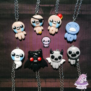 Puede incluir: Una colección de collares con colgantes de personajes hechos a mano. Los colgantes presentan varias figuras de dibujos animados en diferentes colores, incluyendo azul, negro, blanco y beige, con collares de cadena plateada. Los personajes tienen ojos grandes y están llorando.