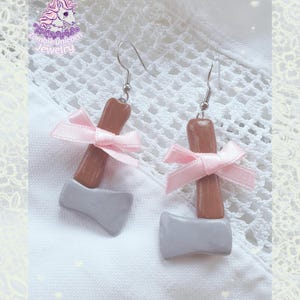 May include: A pair of novelty earrings featuring a gray axe head and a brown handle, accented with a pink ribbon bow. The earrings have silver-colored hooks. The image includes the text "Zombie Unicorn Jewelry".