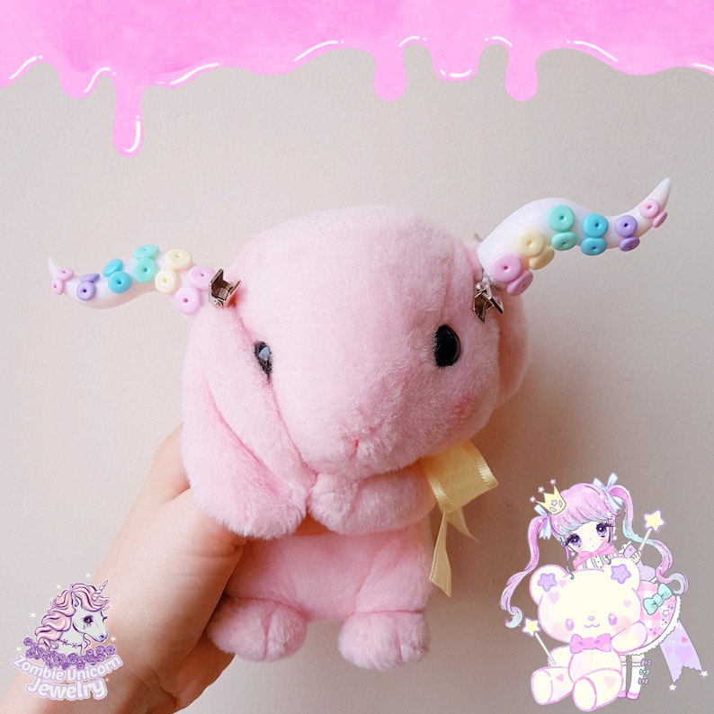 May include: Pink plush bunny with rainbow and white beaded unicorn horns. The bunny is holding a yellow ribbon. The bunny is being held by a hand.