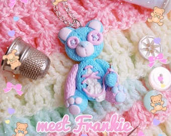 May include: A handmade blue teddy bear pendant necklace with pink accents, button eyes, and a silver chain. The bear is on a colorful, textured background with the text "meet Frankie".