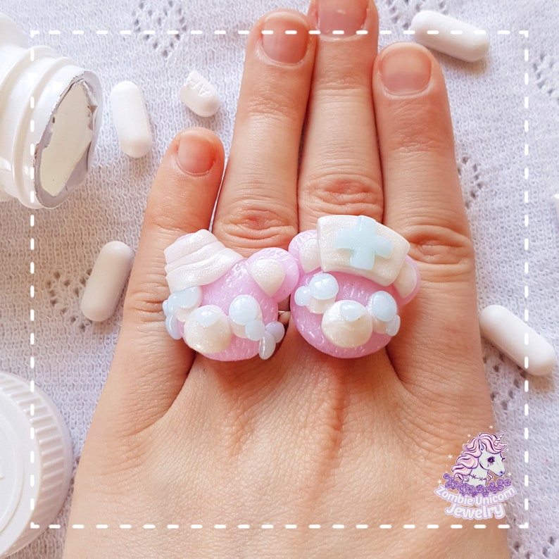 Two pastel pink rings depicting teddy bear heads. One nurse and one patient with bandage on its head. In the background a hand wearing rings and headache pills.