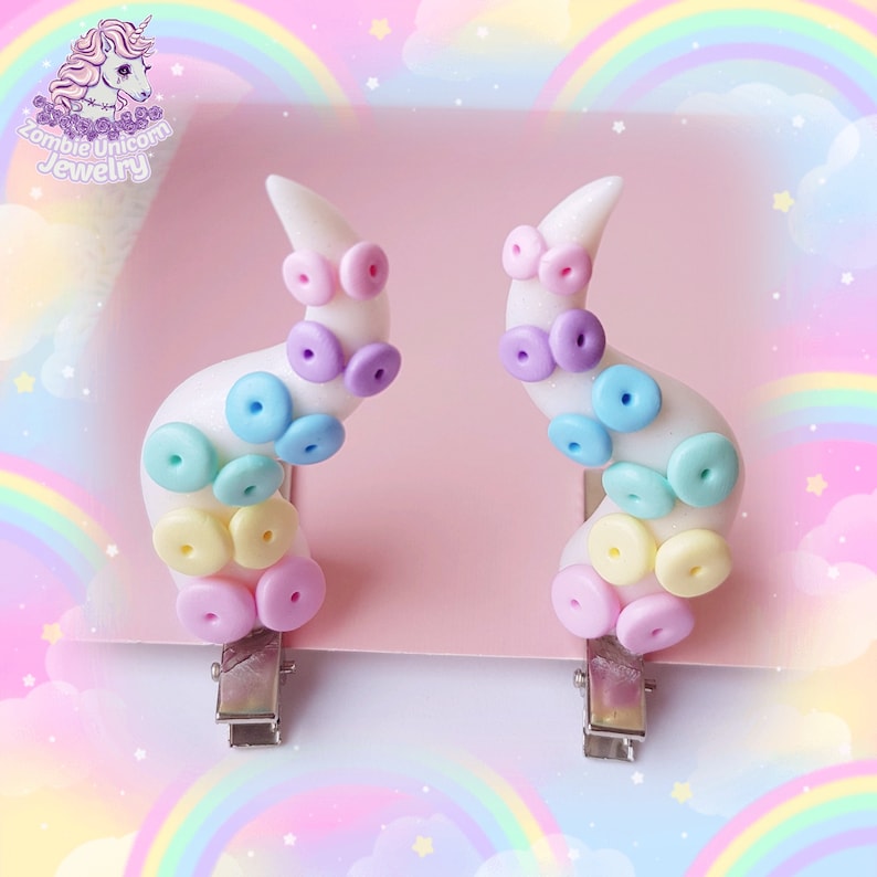 May include: A pair of white hair clips shaped like an octopus tentacle with pastel coloured buttons. The buttons are in a rainbow pattern of pink, yellow, light blue, purple, and light green.