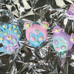 Op de afbeelding: Drie pastelkleurige polymer clay charms met cartoon-aliengezichten met sterren en manen. De charms zijn lichtblauw, roze en paars met gele sterren en een halve maan.