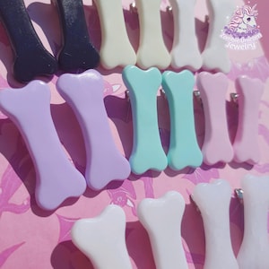 Peut inclure: Collection de barrettes en forme d'os dans diverses couleurs pastel, dont lavande, vert menthe et rose. Les barrettes sont disposées sur une surface rose, avec le logo Zombie Unicorn Jewelry visible en haut à droite.