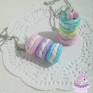 Puede incluir: Dos collares de macarons de color pastel. Un collar presenta una pila de macarons en tonos de azul, amarillo y rosa. El otro collar presenta un solo macaron con un patrón de espiral en tonos de rosa, morado y azul.
