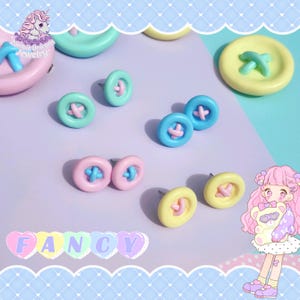 Könnte beinhalten: Knopf-Ohrstecker in Pastellfarben: Rosa, Blau, Gelb und Mintgrün. Jeder Ohrring hat einen kontrastierenden kreuzförmigen Akzent. Das Wort "FANCY" wird in Pastellbuchstaben angezeigt. Ein skurriles Accessoire.