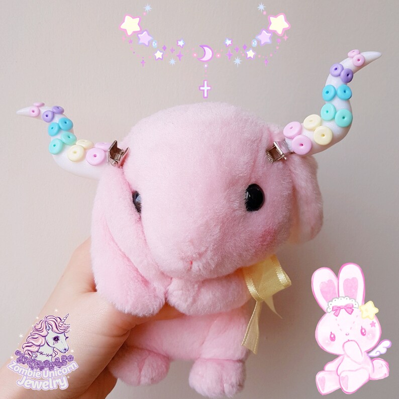 May include: Pink plush bunny with rainbow and white beaded unicorn horns attached with silver clips. The bunny is holding a yellow ribbon. The background is a light pink with a white border and a pattern of stars and a crescent moon.