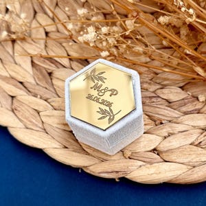 Könnte beinhalten: Eine sechseckige Ringbox mit goldfarbener Oberseite und hellgrauer Samtbasis. Die goldene Oberseite ist mit den Initialen "M&P" und dem Datum "26.06.2026" sowie Blattmotiven graviert. Die Box steht auf einer gewebten Oberfläche.