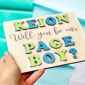以下が含まれることがあります： 青い文字と緑の文字で「Keion Will you be our Page Boy?」と書かれた木製の看板。文字は木目調の背景に貼り付けられている。