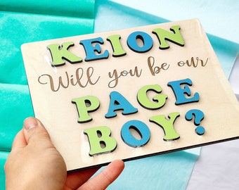 Op de afbeelding: Een houten bord met de tekst "Keion Will you be our Page Boy?" in blauwe en groene letters. De letters zijn bevestigd aan het bord met een houtnerf achtergrond.