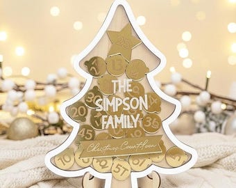 Può includere: Calendario dell'Avvento a forma di albero di Natale in legno, con cornice bianca e dettagli dorati. Presenta una stella e monete d'oro numerate. Il testo "THE SIMPSON FAMILY" e "Christmas Countdown" sono visibili.
