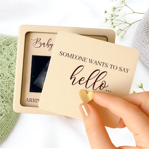 Könnte beinhalten: Eine beige Schwangerschaftsankündigungskarte mit den Worten "Someone wants to say hello..." und einem kleinen goldenen Herzen. Die Karte wird vor einem Rahmen mit dem Wort "Baby" und einem Ultraschallbild gehalten.