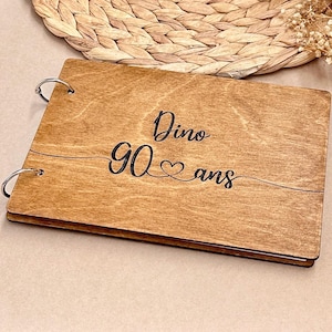 Peut inclure: Un album photo en bois avec reliure à anneaux métalliques. La couverture est gravée des mots "Dino 90 ans" en écriture noire, avec un symbole de cœur. L'album a une finition en grain de bois naturel.