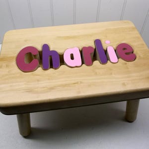 Peut inclure: Un tabouret en bois avec le nom "Charlie" écrit en lettres roses, violettes et marron.