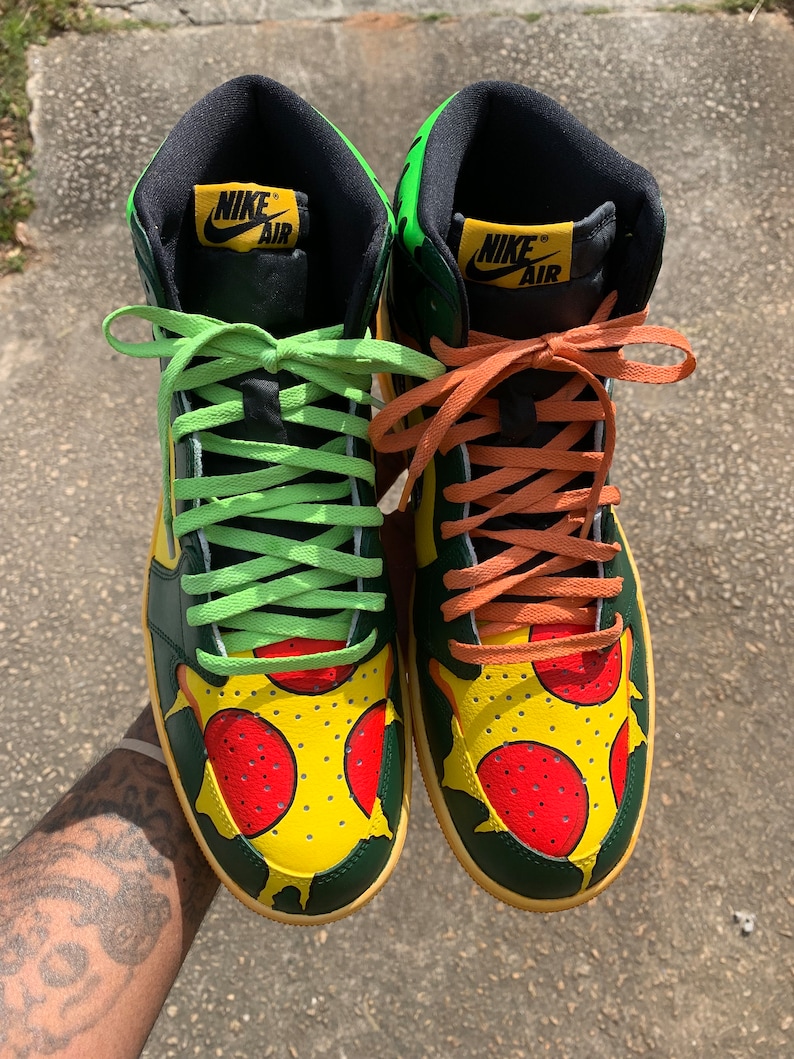 May include: Pair of high-top sneakers with a pizza-themed design. The shoes feature a yellow base with red pepperoni and green accents. The laces are lime green and orange. The Nike Air logo is visible on the tongue.