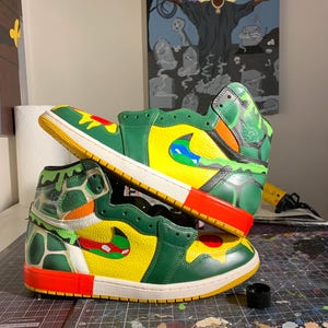 May include: A pair of custom-painted high-top sneakers. The shoes feature a vibrant color scheme with green, yellow, red, and blue accents. The design includes a camouflage pattern on the ankle and a unique Swoosh logo. The soles are white and yellow.