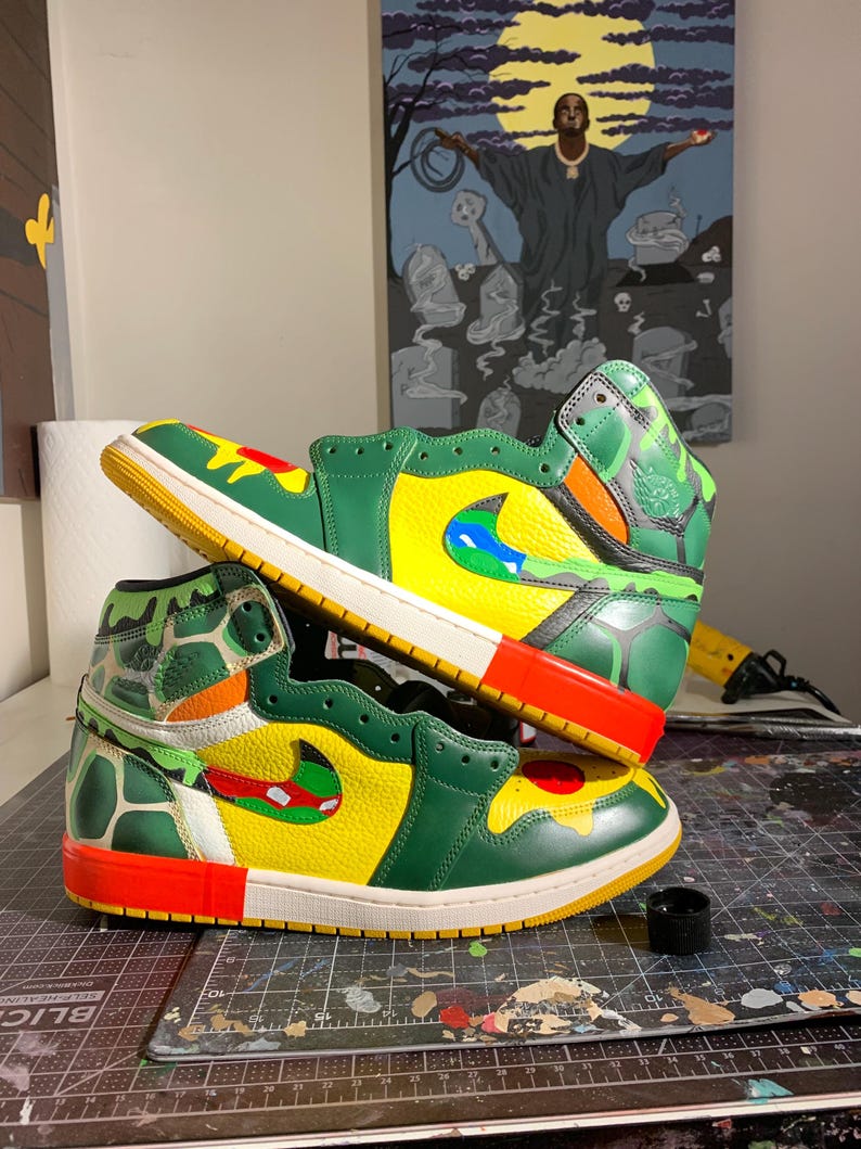 May include: A pair of custom-painted high-top sneakers. The shoes feature a vibrant color scheme with green, yellow, red, and blue accents. The design includes a camouflage pattern on the ankle and a unique Swoosh logo. The soles are white and yellow.