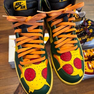 May include: Custom high-top sneakers with a pizza design. The shoes feature a green and yellow base with a pizza slice graphic on the toe box, complete with red pepperoni. Orange laces and a black collar complete the design.