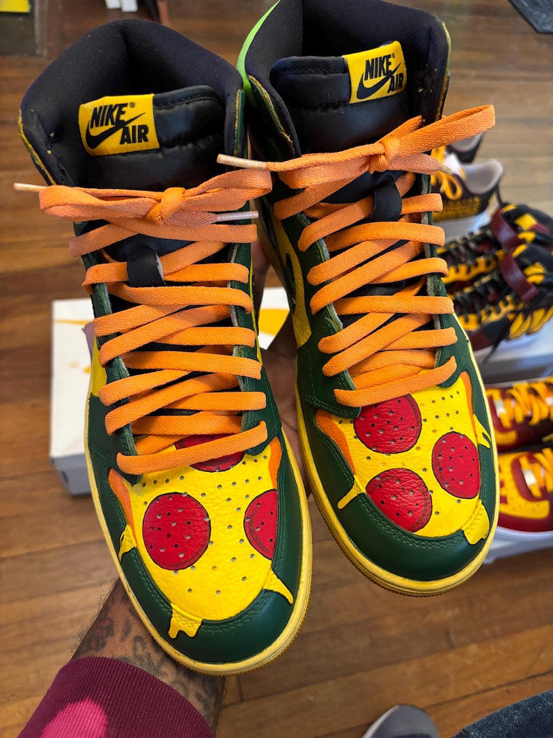May include: Custom high-top sneakers with a pizza design. The shoes feature a green and yellow base with a pizza slice graphic on the toe box, complete with red pepperoni. Orange laces and a black collar complete the design.