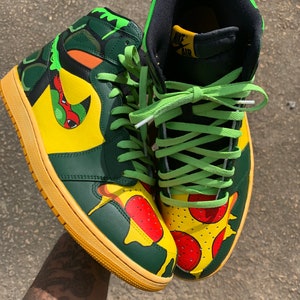 May include: Custom high-top sneakers with a pizza and turtle design. The shoes feature a yellow, green, and orange color scheme with green laces and a yellow sole. The design includes pizza slices and a turtle-like pattern.
