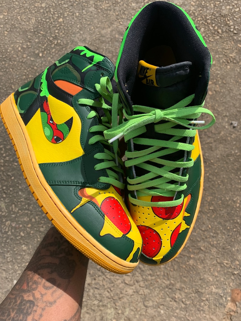 May include: Custom high-top sneakers with a pizza and turtle design. The shoes feature a yellow, green, and orange color scheme with green laces and a yellow sole. The design includes pizza slices and a turtle-like pattern.