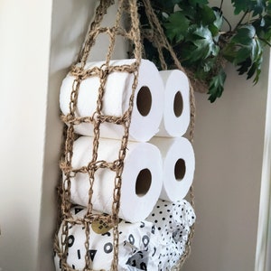 May include: A brown crocheted net hanging from the ceiling holds several rolls of toilet paper. The rolls are stacked on top of each other, with the bottom roll having a black and white polka dot pattern.