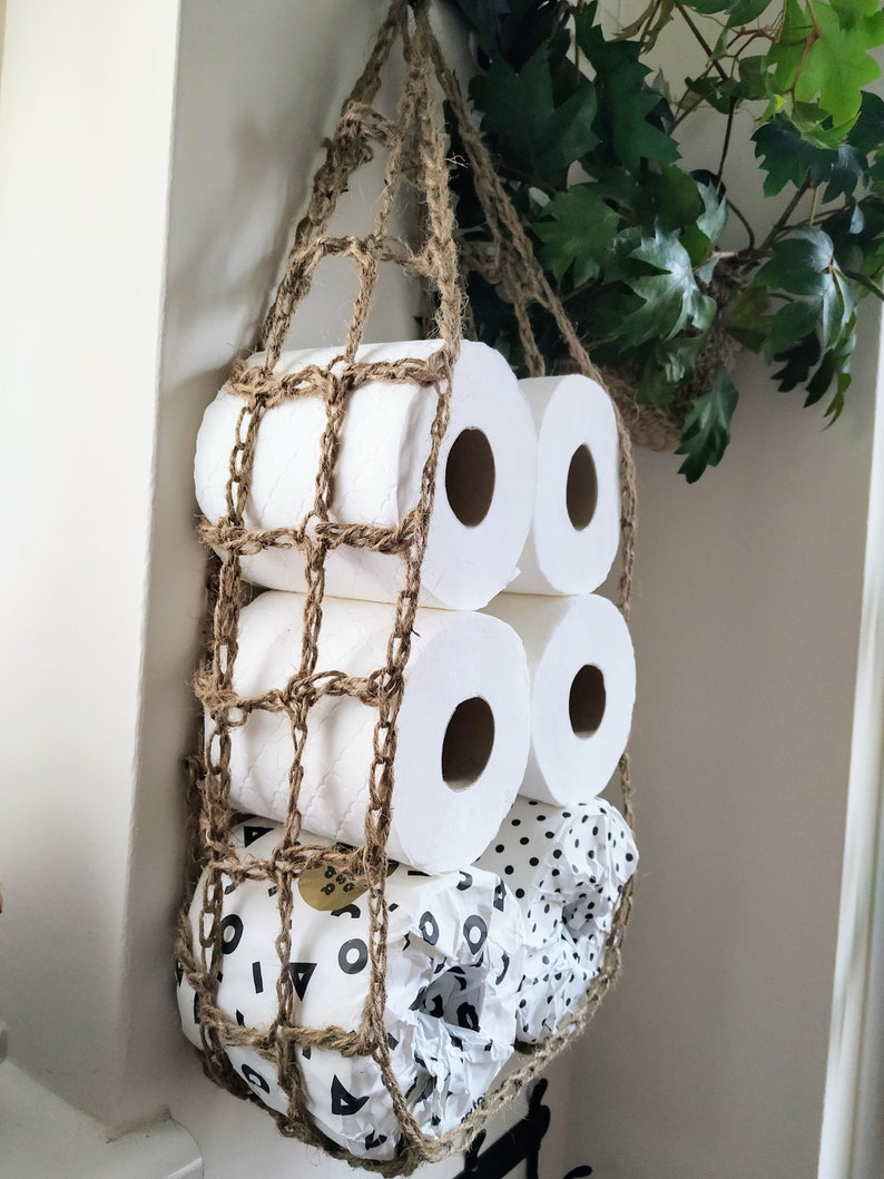 May include: A brown crocheted net hanging from the ceiling holds several rolls of toilet paper. The rolls are stacked on top of each other, with the bottom roll having a black and white polka dot pattern.
