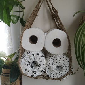 May include: Three rolls of toilet paper are hanging in a woven jute bag. The bag is hung on a hook. Two rolls are white and one roll has a black and white geometric pattern.