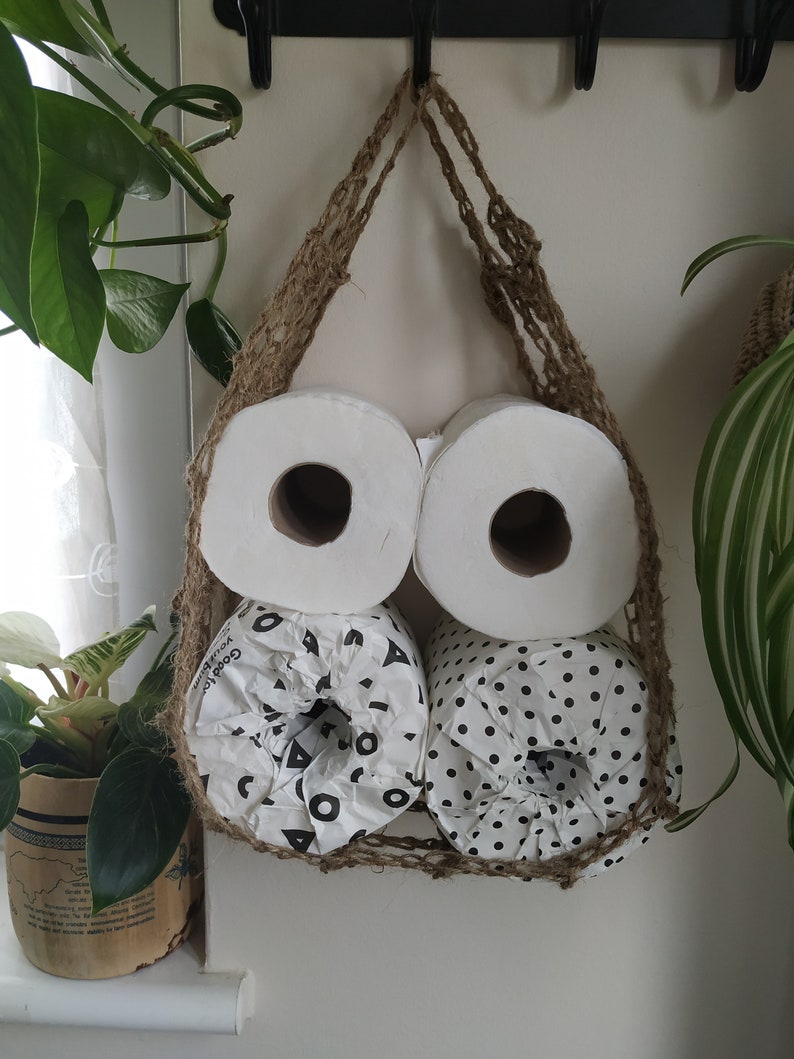 May include: Three rolls of toilet paper are hanging in a woven jute bag. The bag is hung on a hook. Two rolls are white and one roll has a black and white geometric pattern.