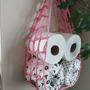 May include: Pink crocheted hanging storage net with three rolls of toilet paper. Two rolls are white and one roll has a black and white geometric pattern.