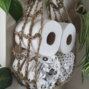 May include: Two rolls of toilet paper and one roll of paper towels are stored in a hanging brown crocheted net bag. The paper towels are wrapped in white paper with black polka dots and the text "Good  Pulp,  Your  World,  Greener  Toilet  Paper" is visible.
