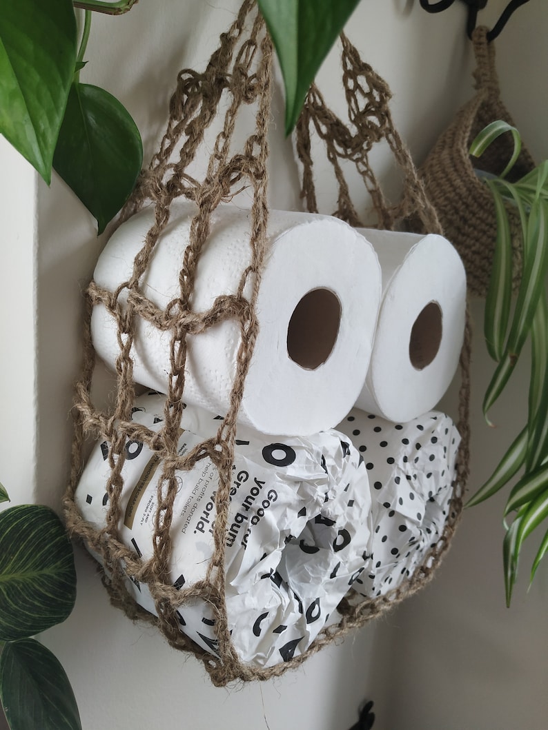 May include: Two rolls of toilet paper and one roll of paper towels are stored in a hanging brown crocheted net bag. The paper towels are wrapped in white paper with black polka dots and the text "Good  Pulp,  Your  World,  Greener  Toilet  Paper" is visible.